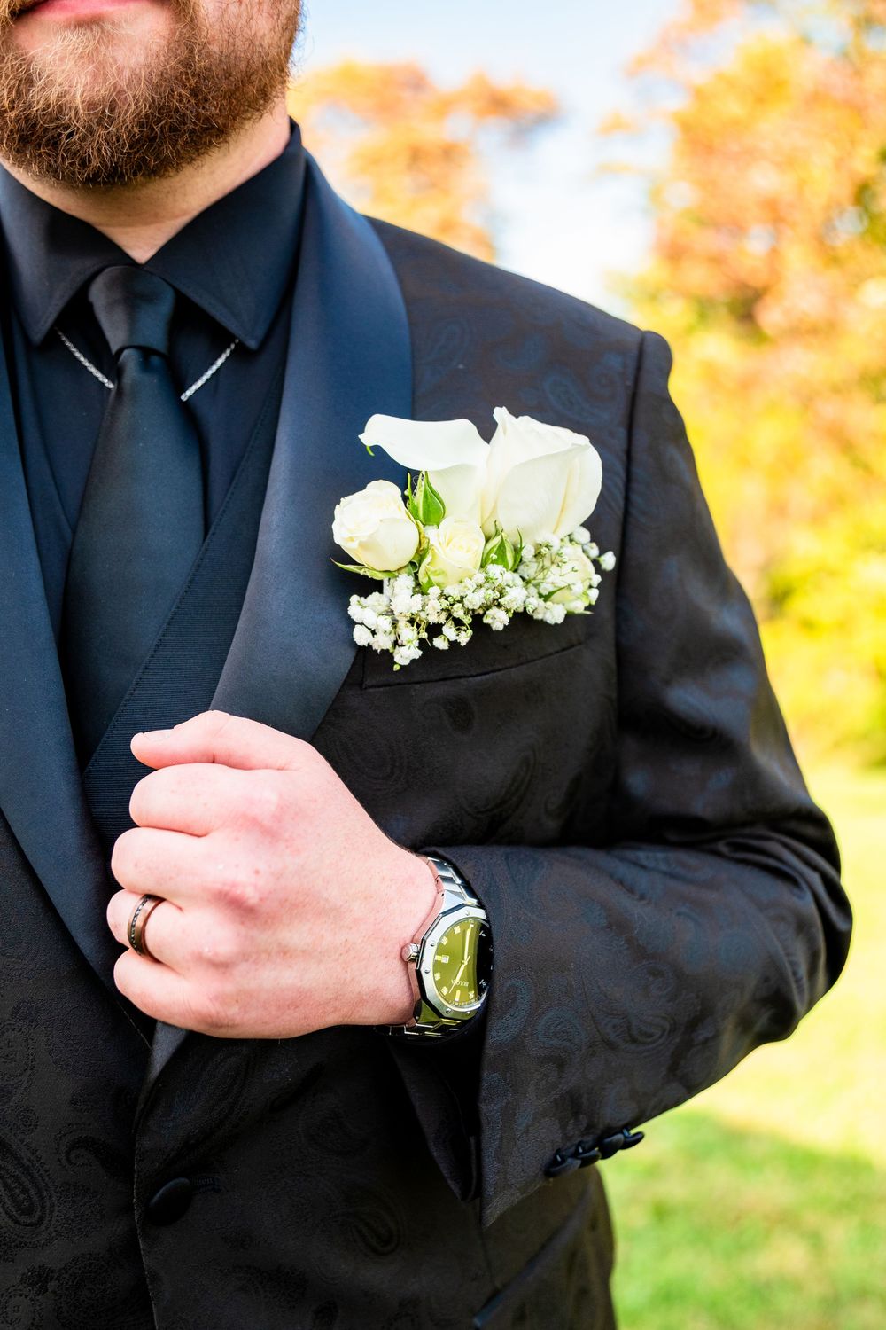 Groom in black tuxedo with white rose boutonniere, wearing a watch and ring, with autumn foliage background.