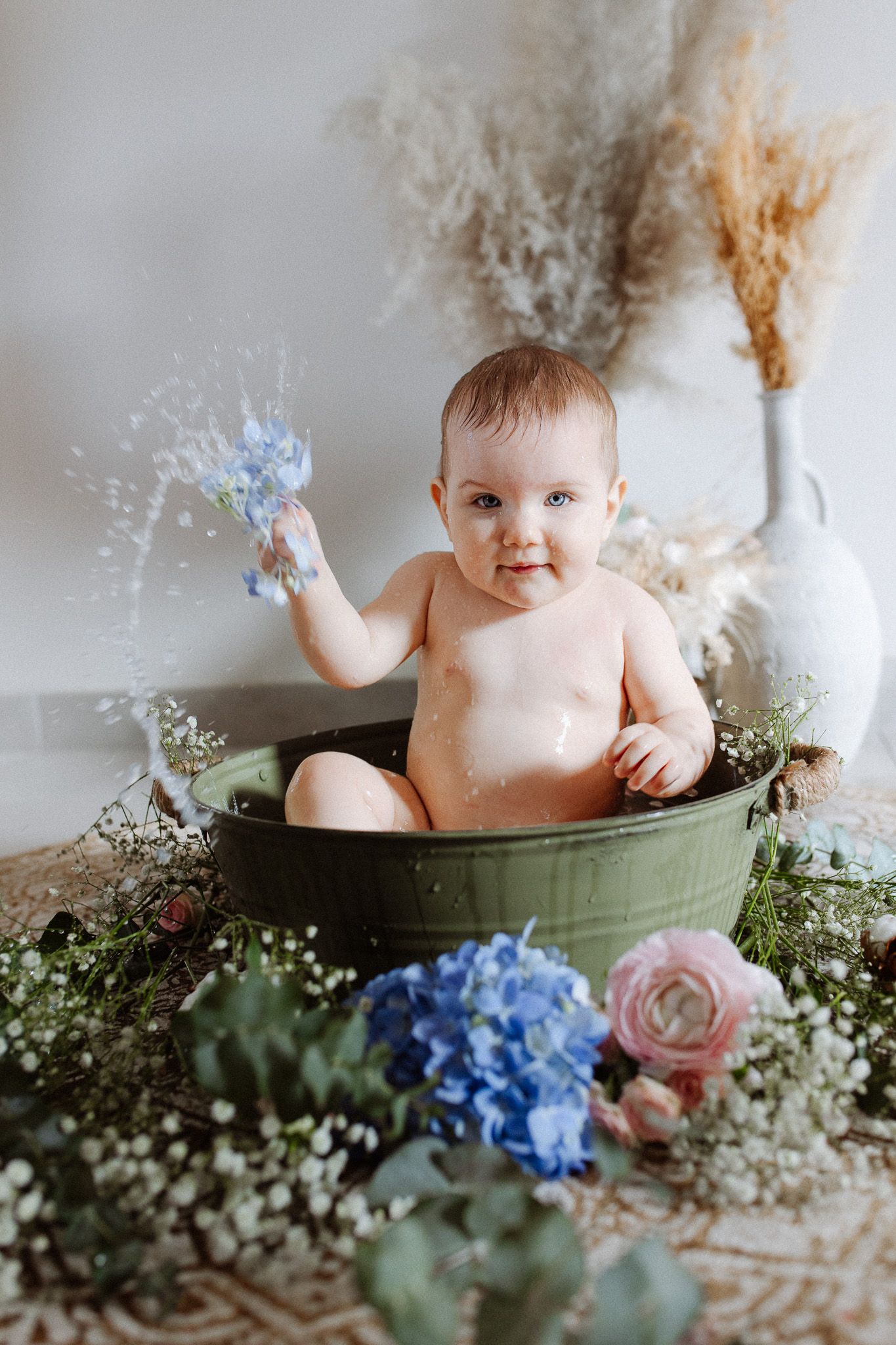 Un bébé assis dans une baignoire verte, entouré d’hortensias bleus et de fleurs blanches, éclaboussant l’eau dans un environnement lumineux.