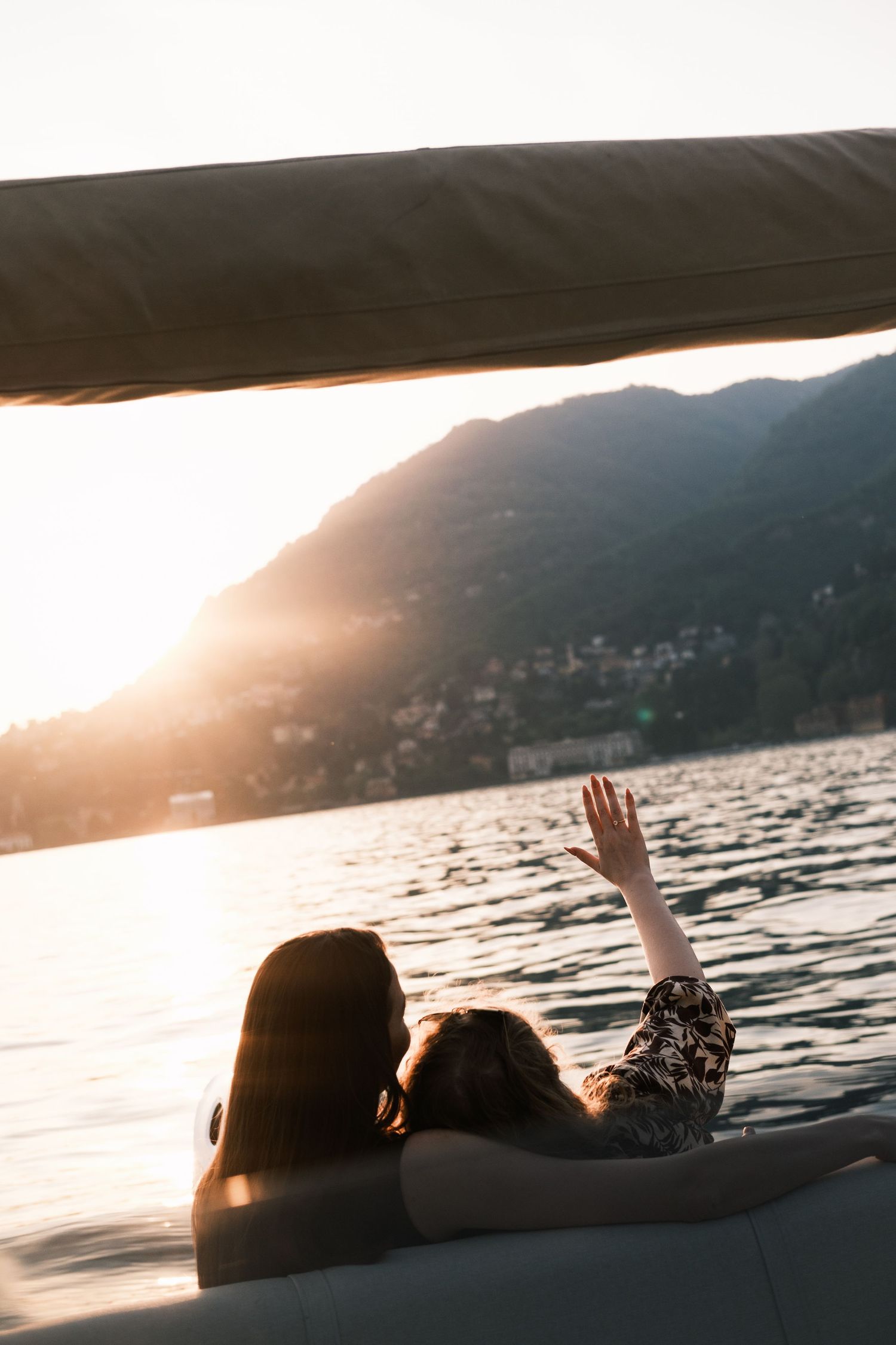 Two women relaxing on a boat at golden sunset over a mountain lake, one raising her hand toward the scenic waterfront.