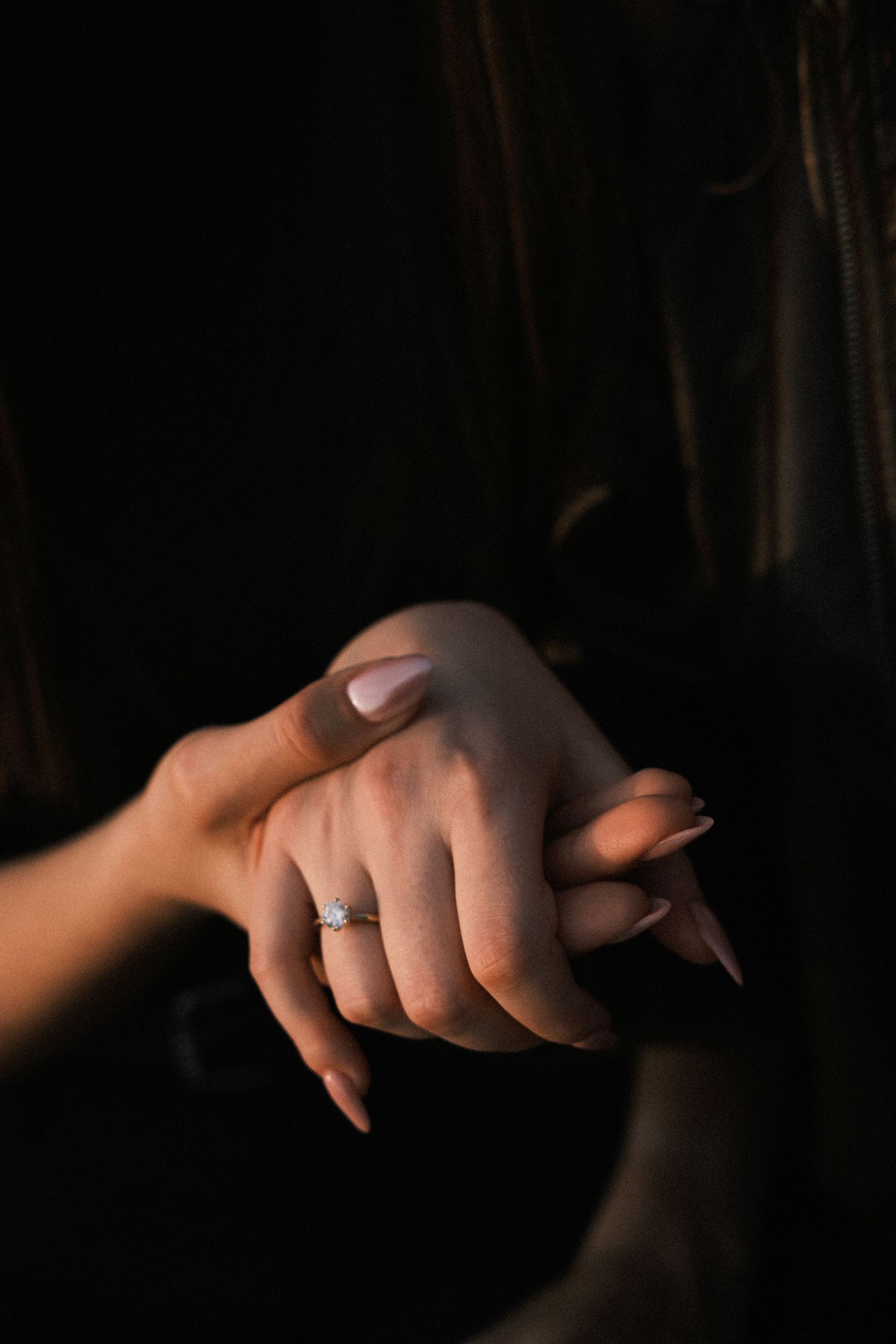 Close-up of a woman's hands with nude manicure wearing a delicate diamond engagement ring against dark background.
