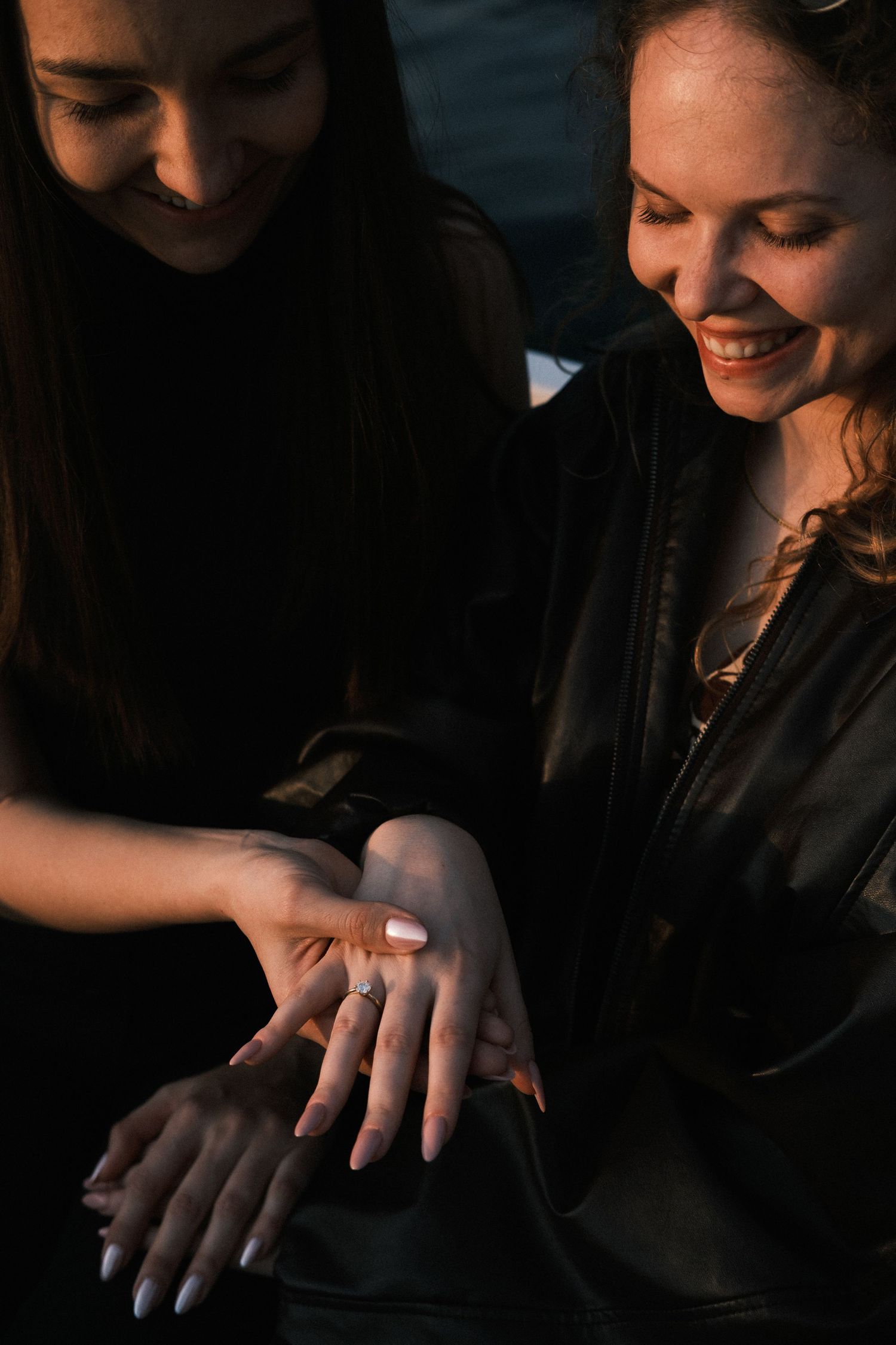 Two smiling women in black outfits admiring an engagement ring on a finger in warm evening light.