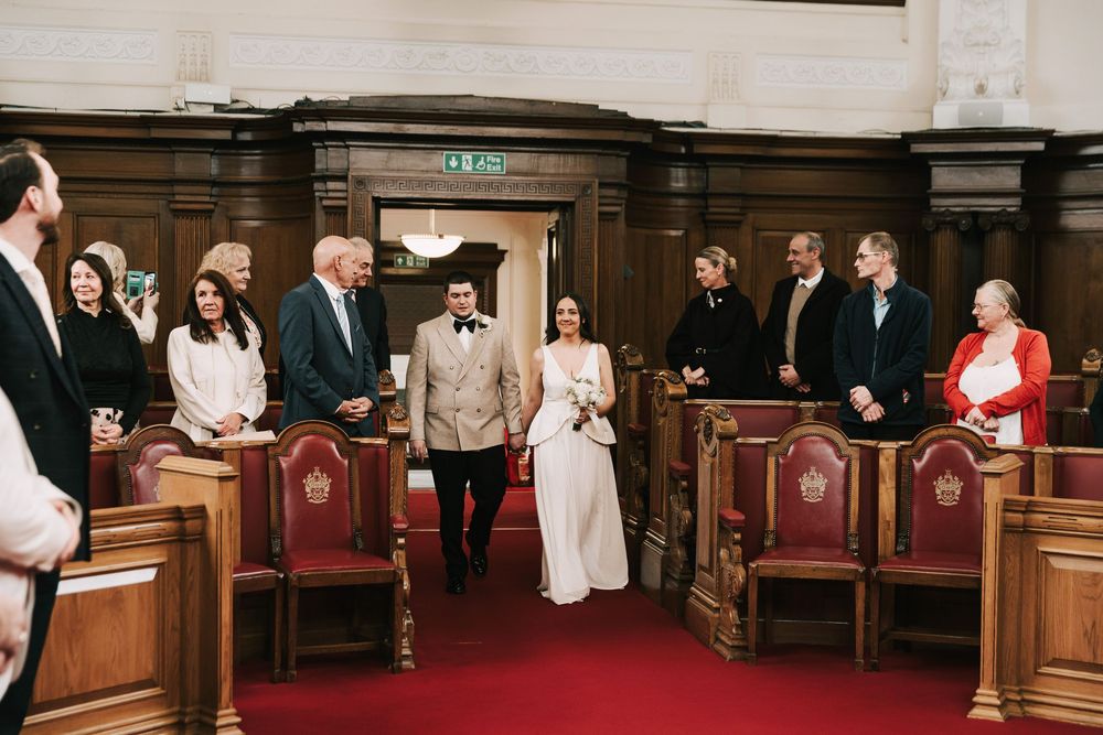 Bride and groom walking down the aisle in an elegant wood-paneled wedding ceremony hall with red chairs.