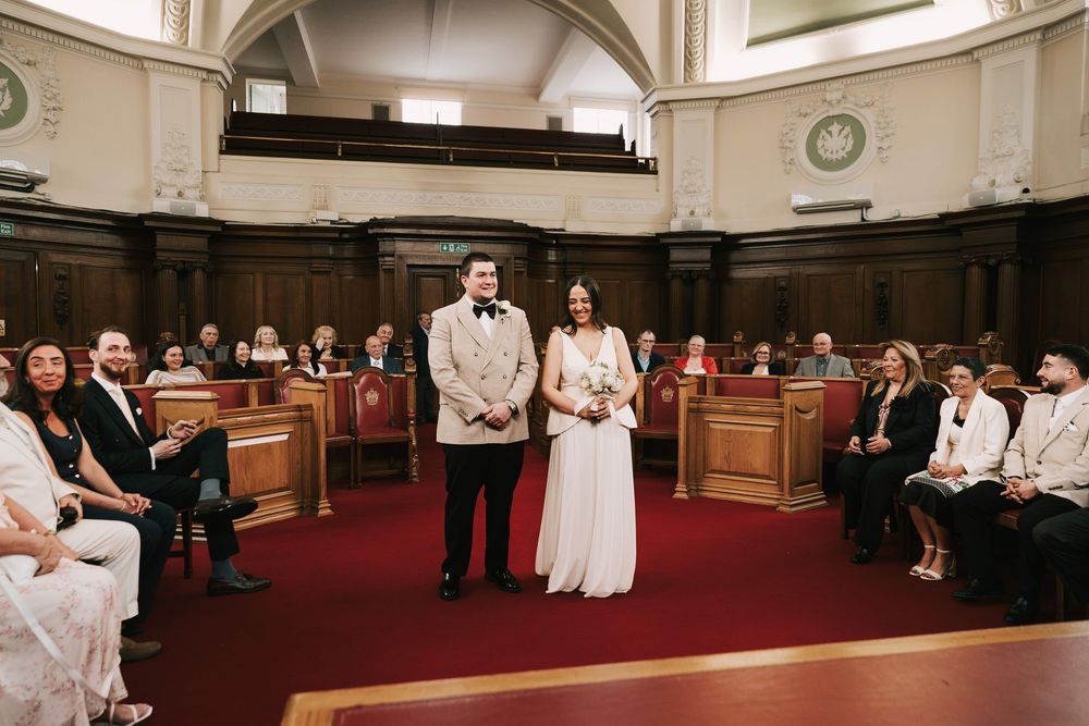 Bride and groom stand together during intimate courthouse wedding ceremony with guests seated in wooden pews.