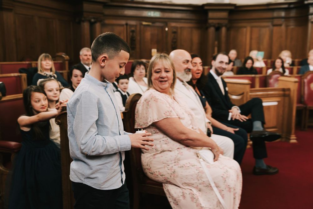 A boy escorts a woman in a pink lace dress to her seat at a wedding ceremony in a classic wood-paneled venue.