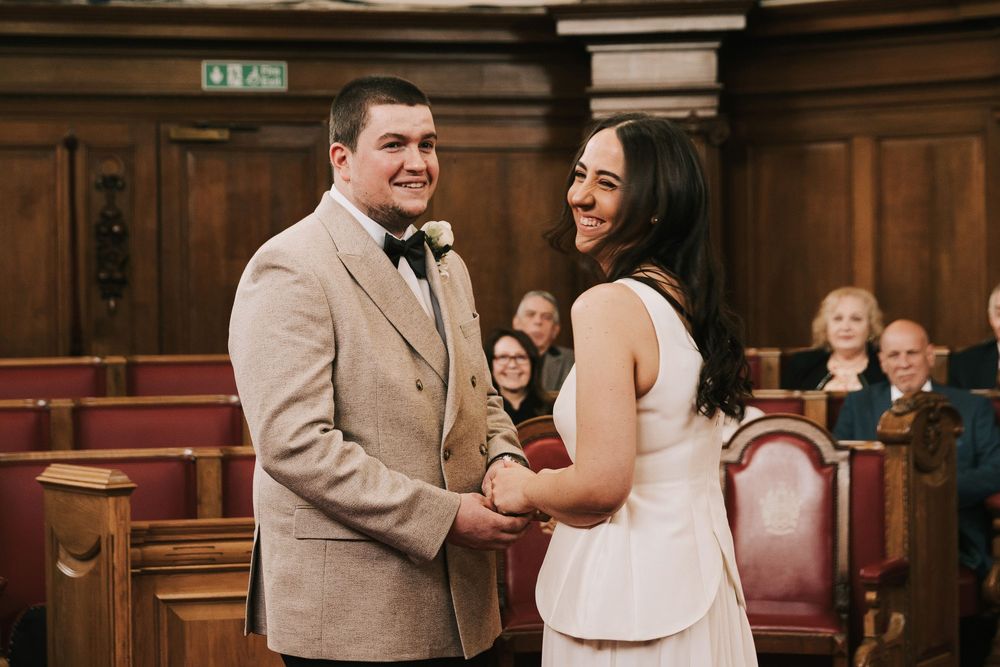 Couple holding hands and smiling at wedding ceremony in wood-paneled room with guests seated behind them.