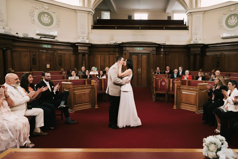 Newlyweds share first kiss as married couple during civil ceremony in grand wood-paneled hall while guests applaud in celebration.