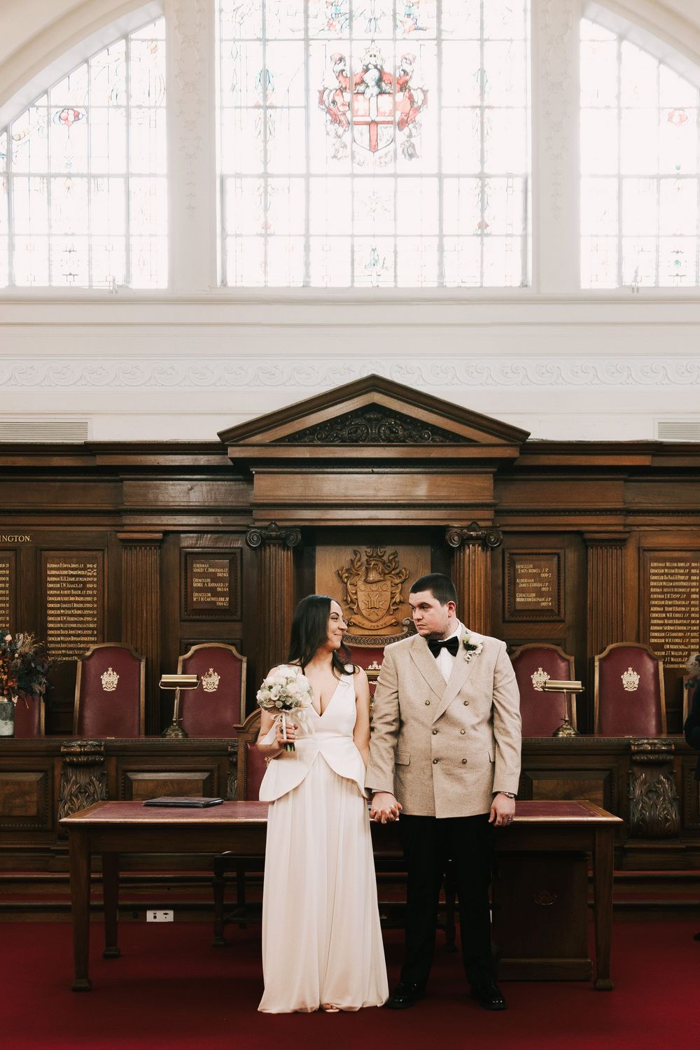 Bride and groom pose before ornate wooden council chamber with stained glass coat of arms at a registry office wedding.