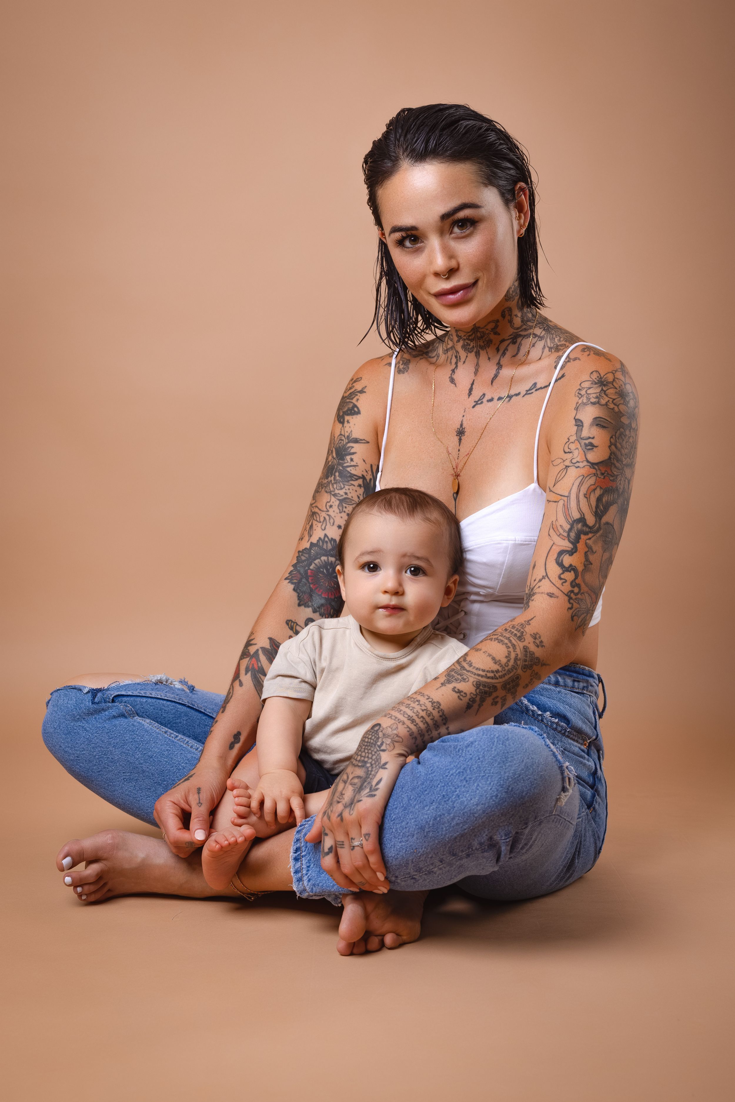 A parent with colorful tattoos sits cross-legged on a beige backdrop while holding a baby in casual clothing.