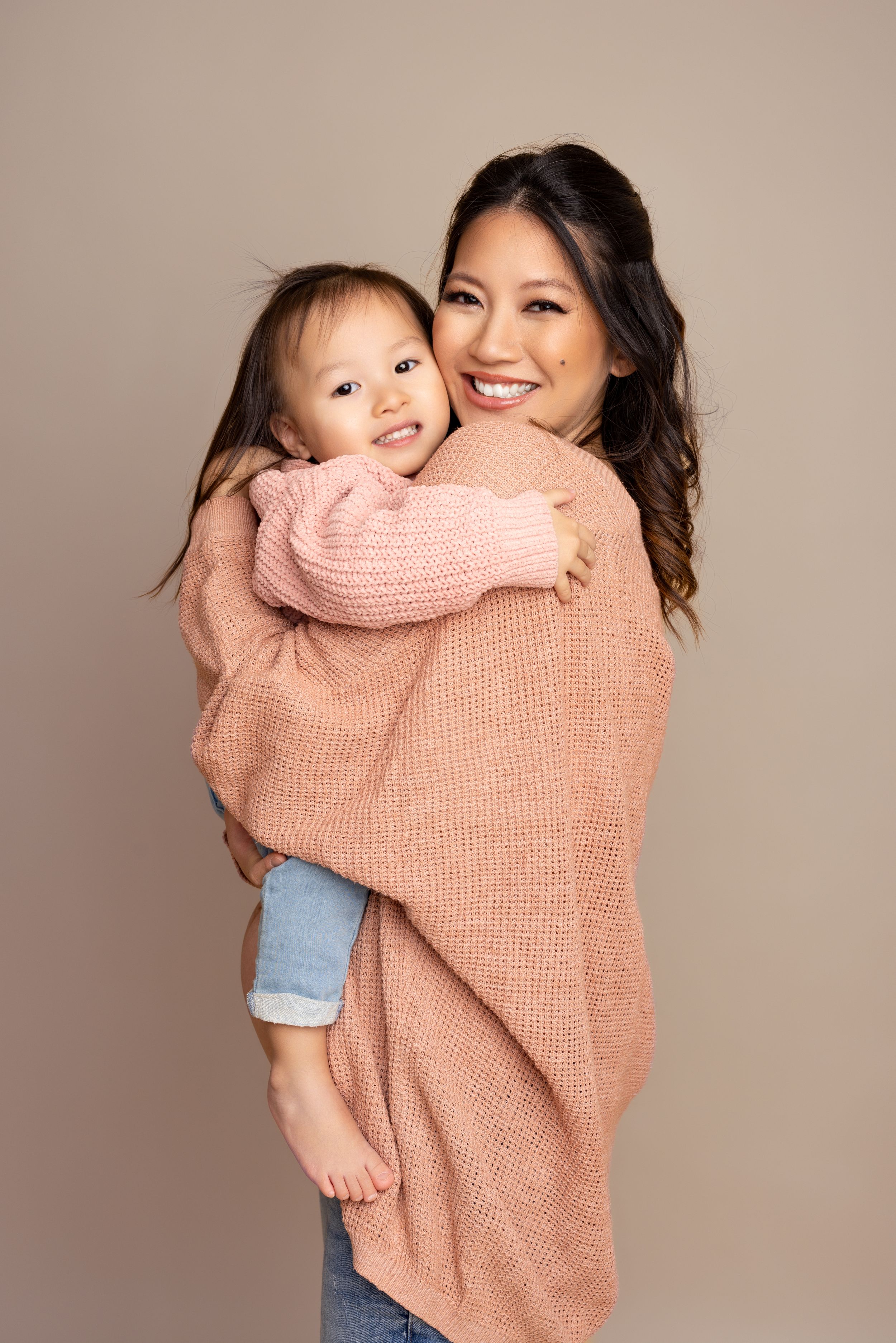 Parent wearing pink sweater and jeans embracing small child in matching outfit against neutral background.