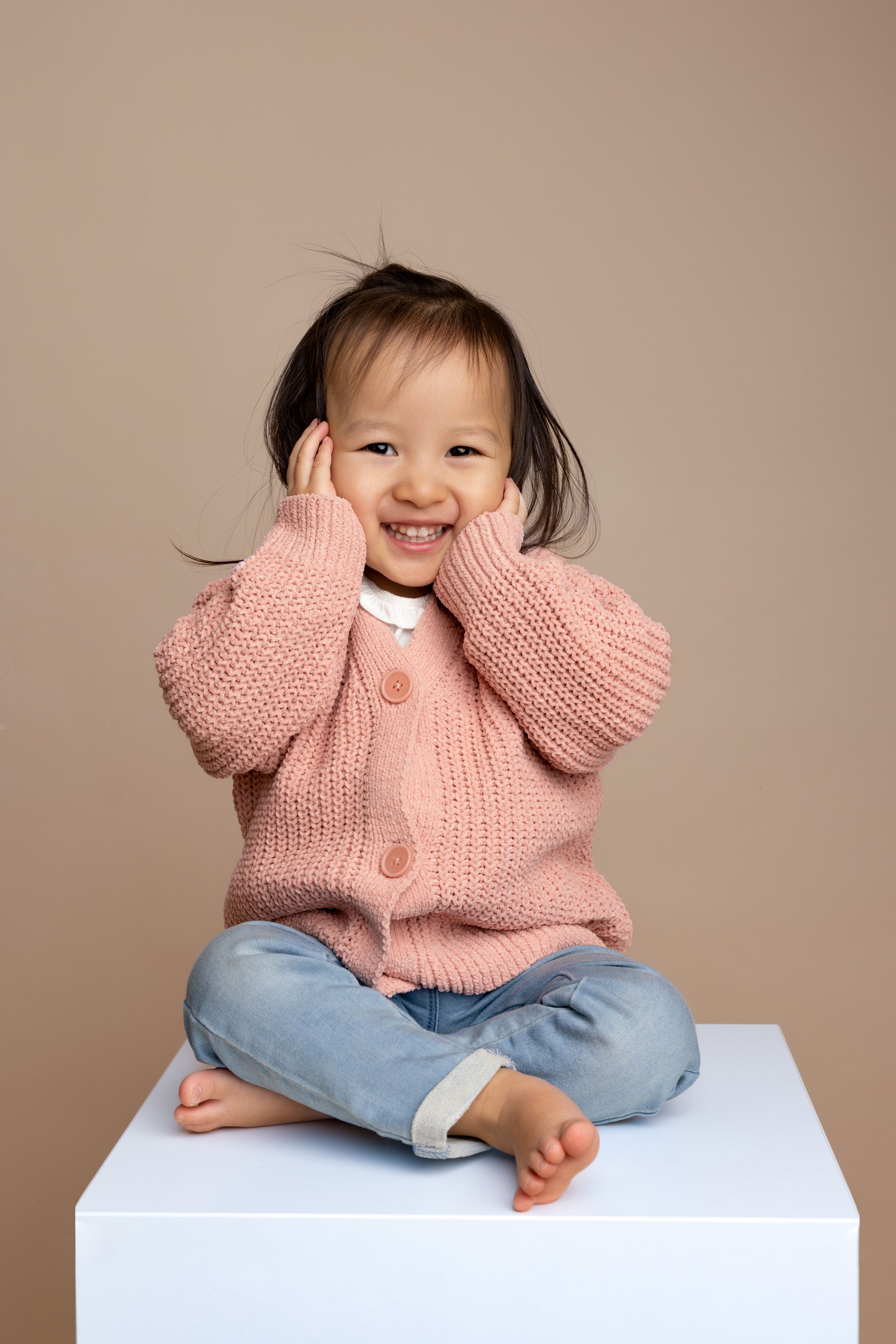 Young child wearing a pink knit sweater and jeans sitting on a white cube while playfully covering their face.