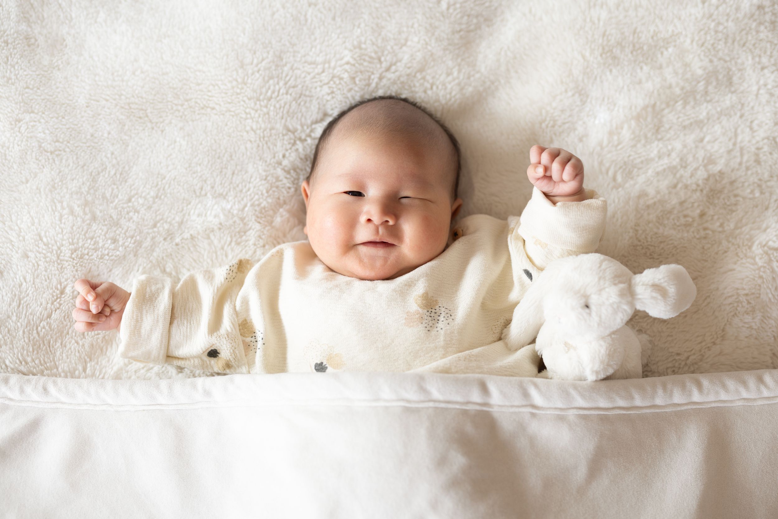 Peaceful sleeping baby in cream outfit with stuffed toy on white bedding.