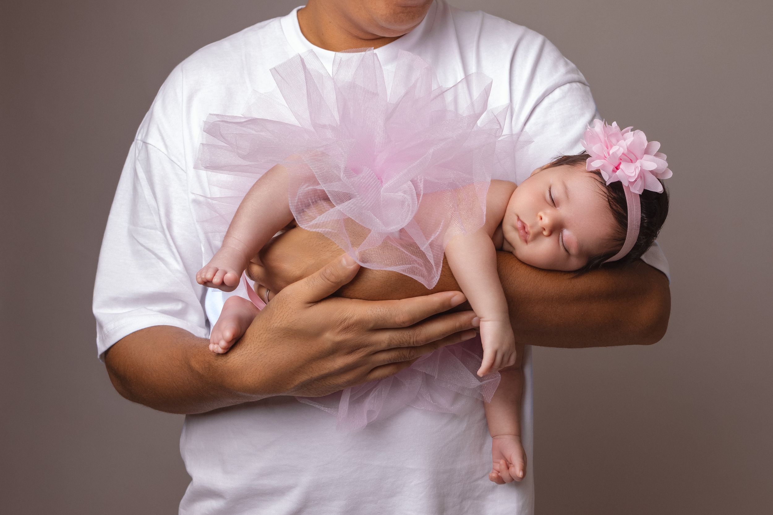 A sleeping baby in pink tutu is gently cradled in parent's arms wearing white t-shirt.