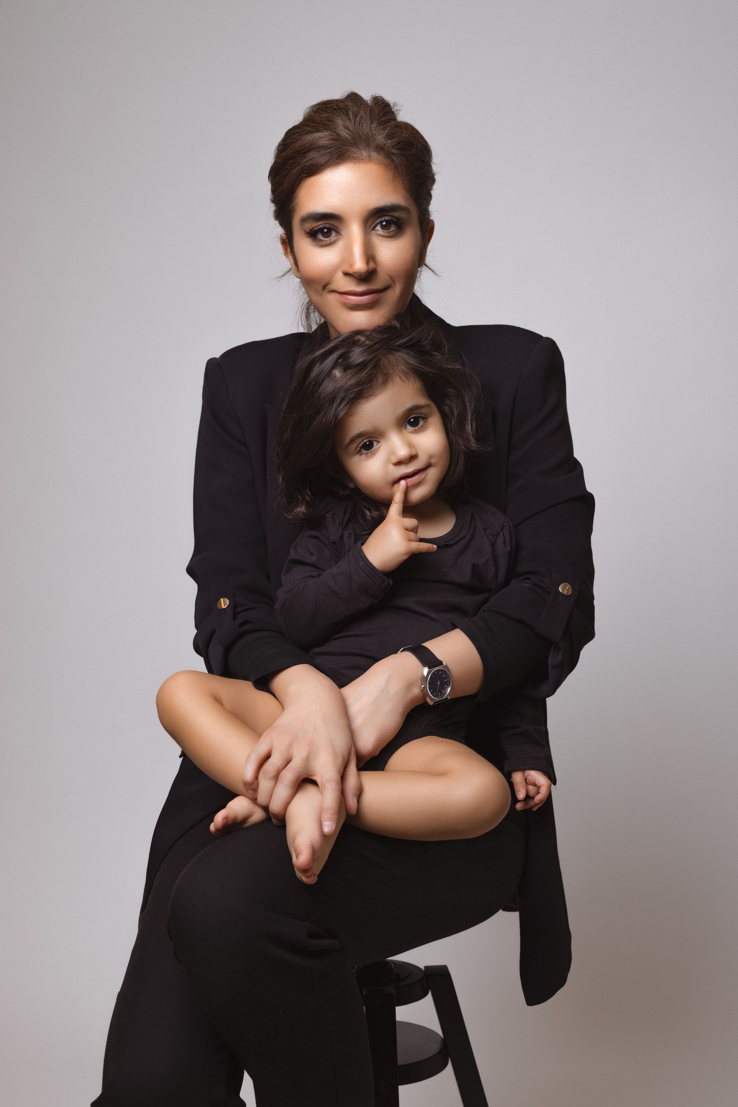 Portrait of two people in matching black outfits sitting together on a stool against gray background.