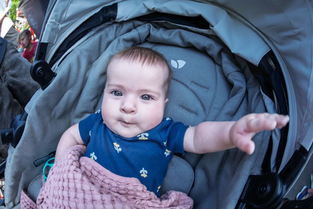 Cute baby in a gray stroller wearing a navy onesie, reaching out with one hand, wrapped in a pink blanket.