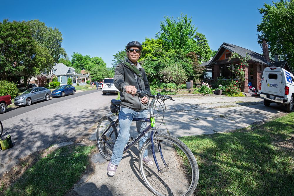 Man in helmet poses with bicycle on sunny residential street lined with trees and parked cars.