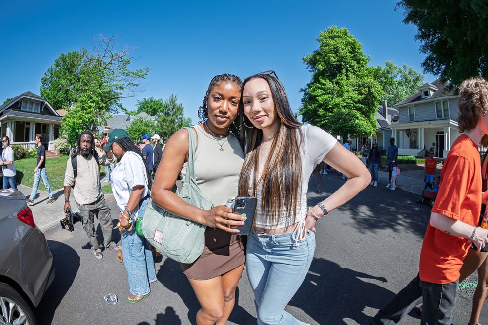 Two young women smile together on a sunny street during an outdoor community event with people gathered in the background.