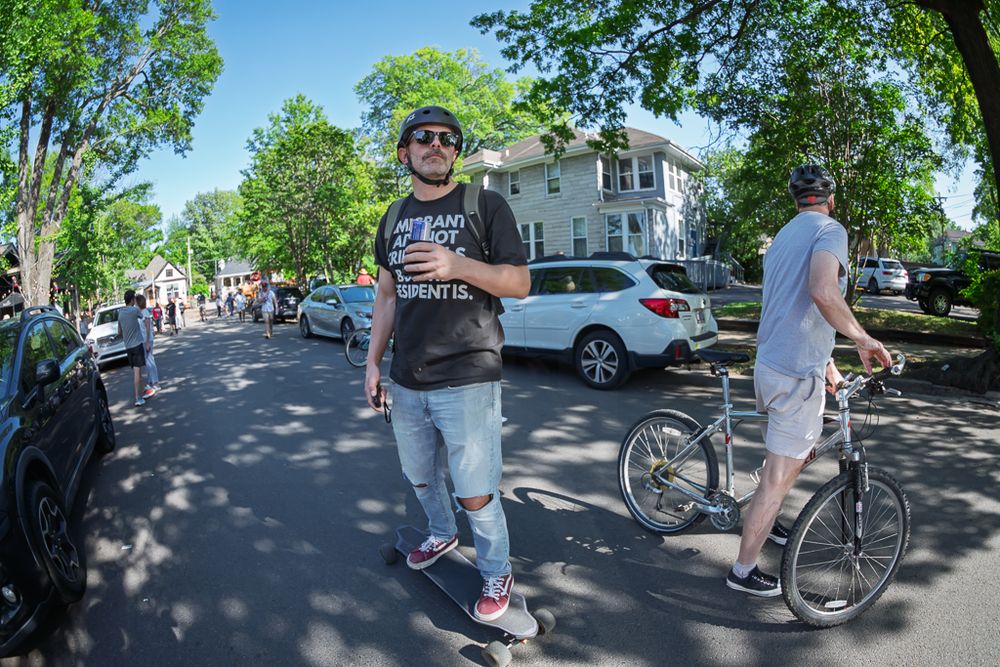 Man skateboarding on a tree-lined street during a sunny outdoor community event with cyclists nearby.