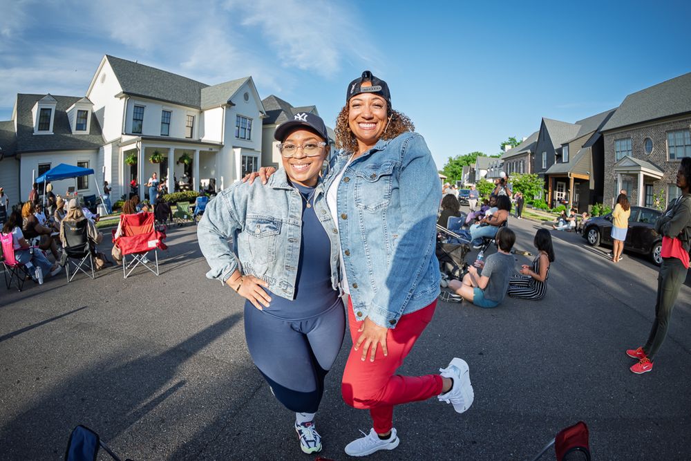 Two women pose together smiling on a street during an outdoor community event on a sunny day.