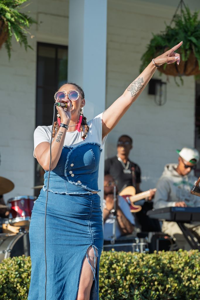 Female singer performing outdoors in denim overalls pointing while band plays behind her on sunny day.
