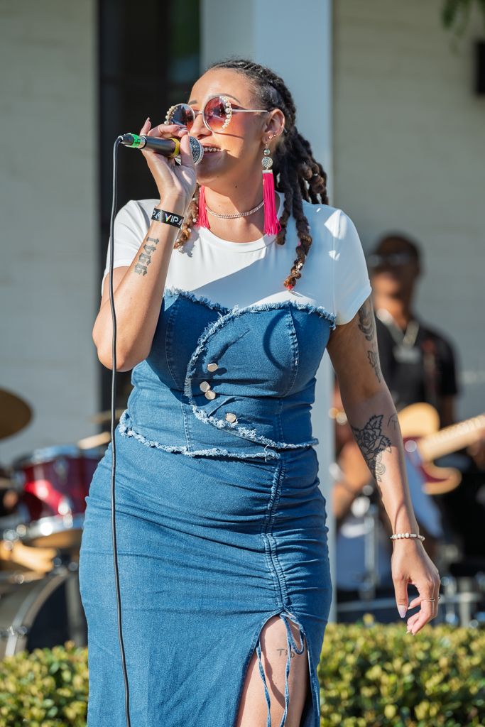Female singer in denim outfit performs outdoors with microphone on a sunny day.