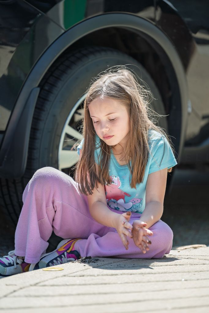 Young girl with long hair sits cross-legged on pavement near a car tire, looking at her hands.