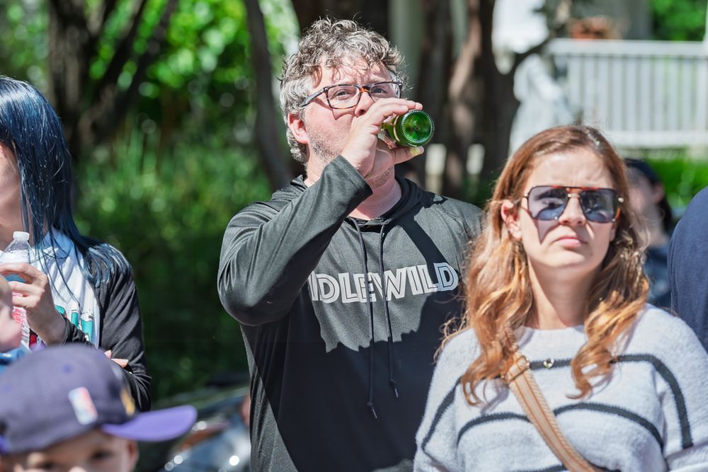 Man drinking from a green bottle at an outdoor event while a woman in sunglasses stands nearby.