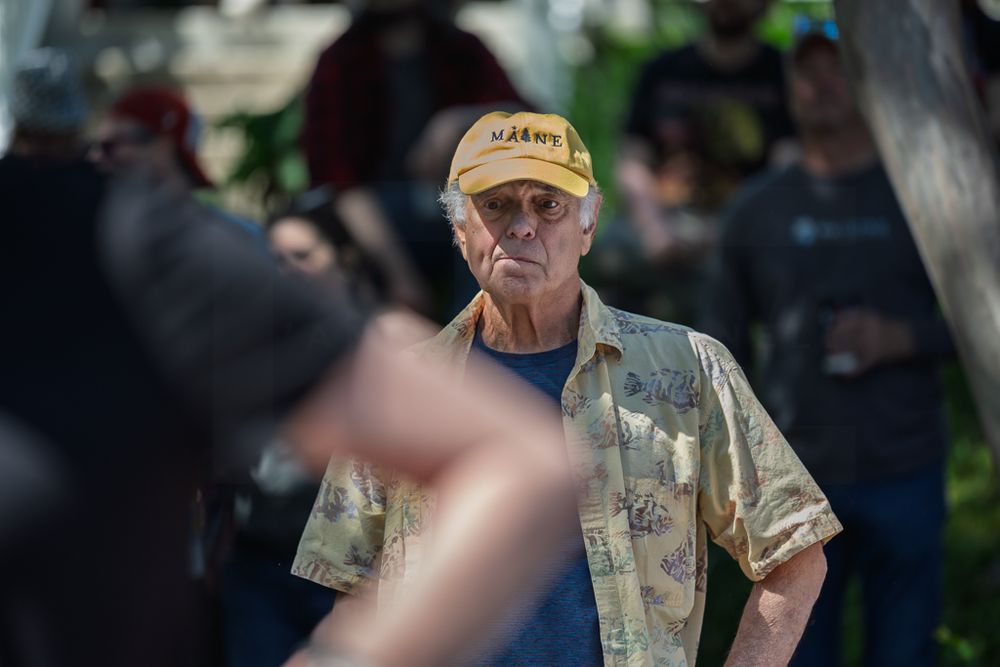 Elderly man in yellow US Army cap and floral shirt standing outdoors at a crowded event.