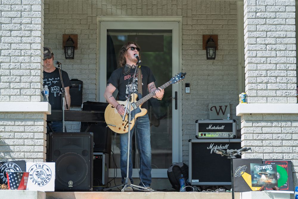 A guitarist performs live music on a porch stage with Marshall amplifiers during an outdoor neighborhood event.
