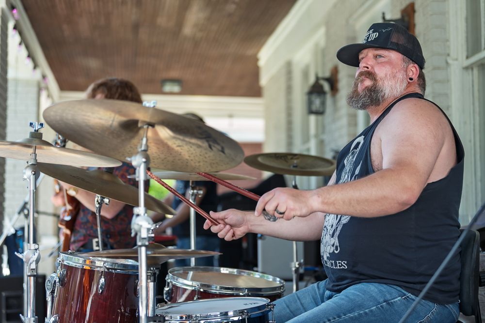 Heavyset male drummer plays cymbals on a covered porch during an outdoor performance.