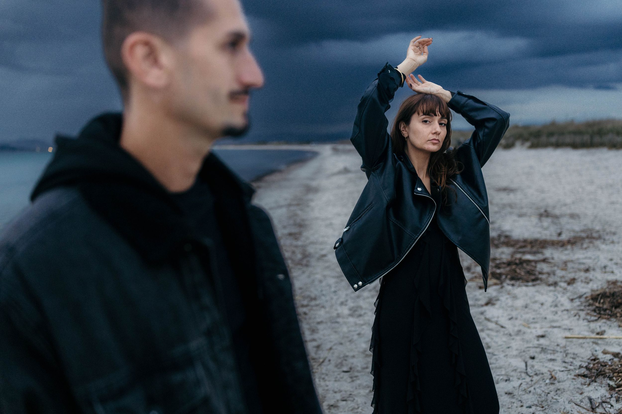 A man and woman pose on a stormy beach, she raises her arms gracefully in a dark outfit under dramatic cloudy skies.