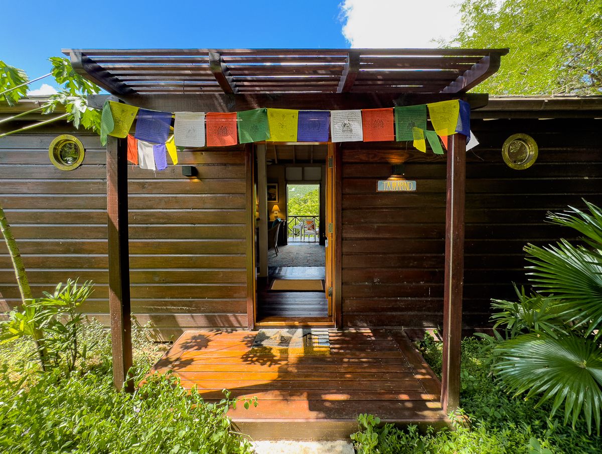 Colorful Tibetan prayer flags hang over a wooden entrance to a tropical treehouse retreat surrounded by lush greenery.