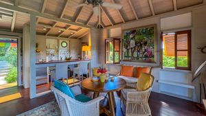 Charming Caribbean cottage interior with wicker chairs, round dining table, vaulted ceiling, and vibrant tropical artwork on the wall.