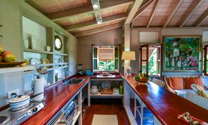 Cozy tropical kitchen with red countertops, wood ceiling, open windows, and colorful artwork in a charming cottage home.