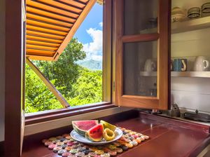 Tropical fruit platter with watermelon and avocado on a kitchen counter beside an open wooden window overlooking lush green mountains.