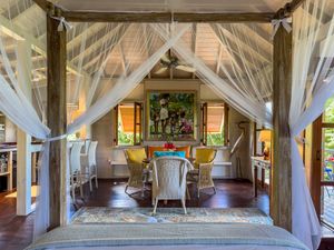 Elegant four-poster bed with sheer canopy drapes in a tropical villa bedroom with dining area and lush artwork.