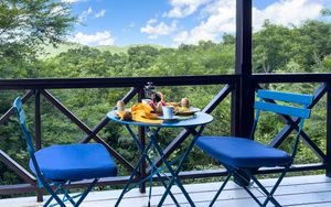 Blue bistro chairs and table on a wooden balcony overlooking a lush tropical forest canopy under a bright blue sky.