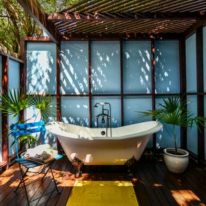 Luxurious outdoor clawfoot bathtub on a wooden deck surrounded by tropical plants and blue panels.