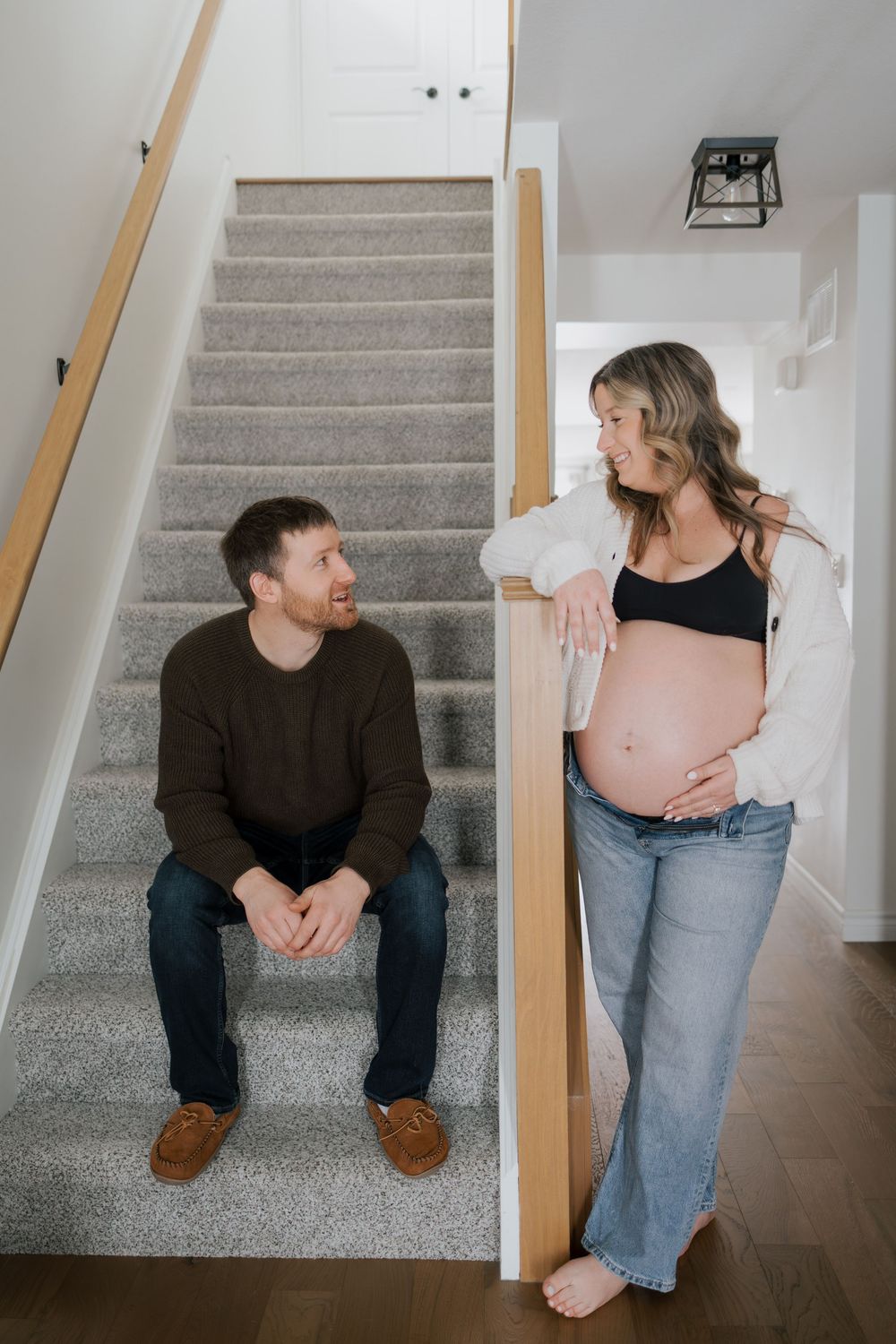 Expecting couple sharing a smile near staircase, man seated on steps, pregnant woman leaning on railing.