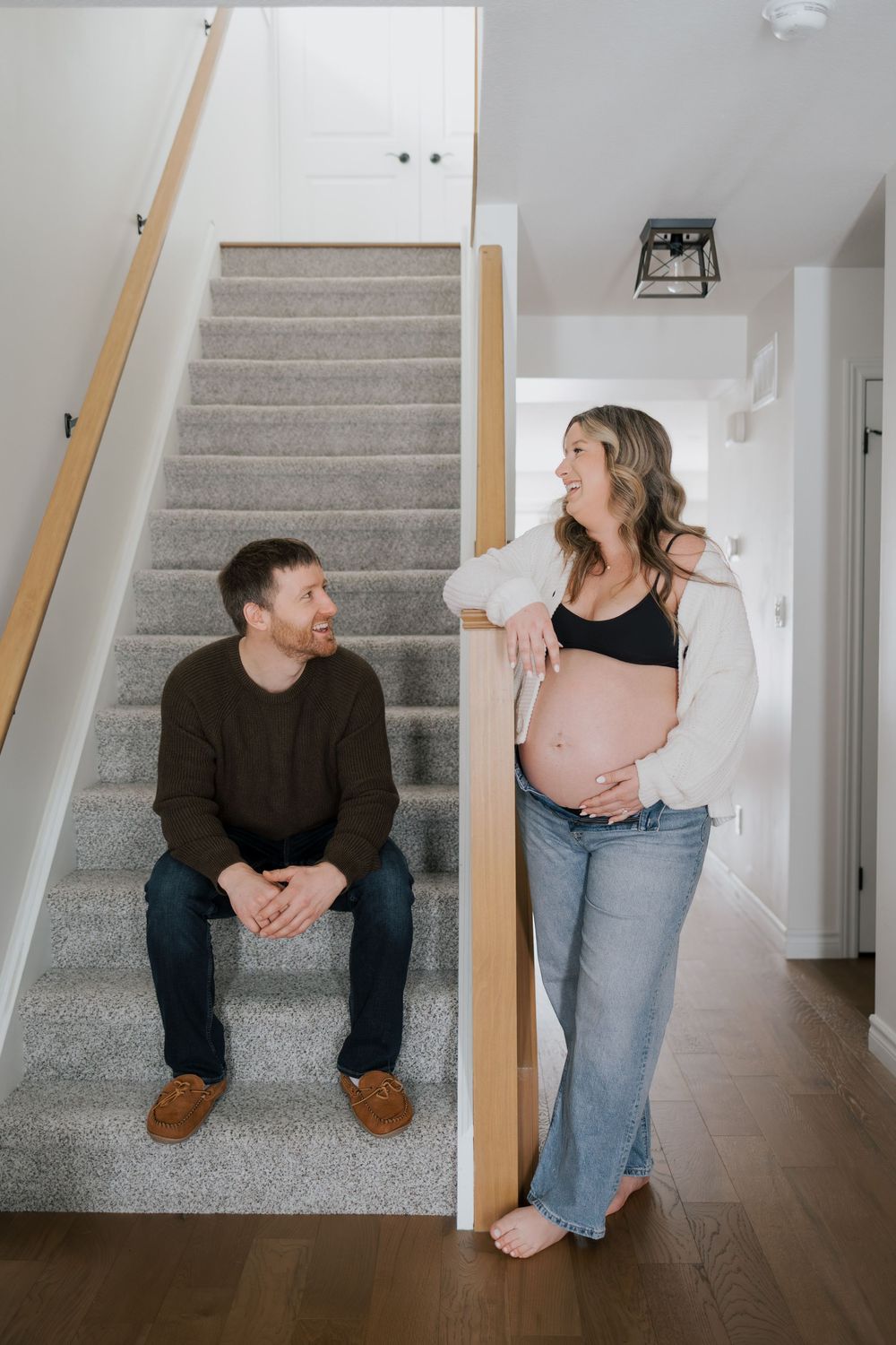 Happy couple at home staircase, pregnant woman in white cardigan holding baby bump, partner seated smiling.