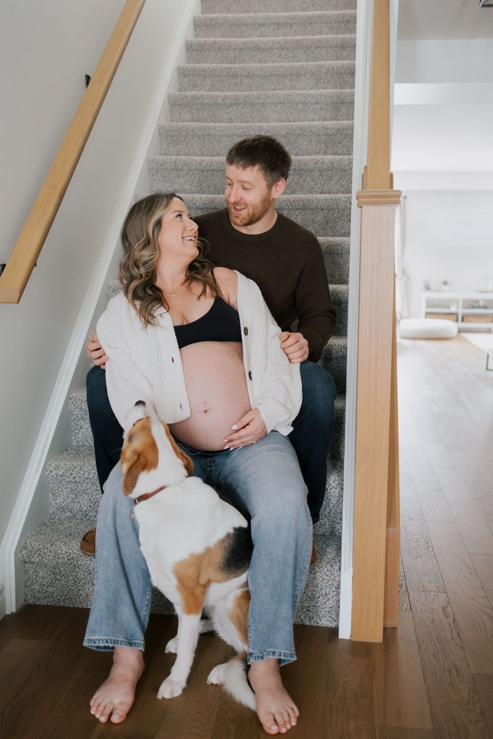 Smiling couple sitting together on home staircase with beagle dog, pregnant woman cradling her baby bump.