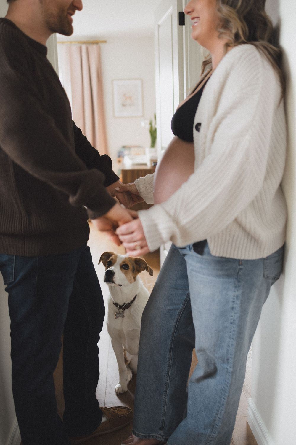 A smiling pregnant couple holds hands in a hallway while their dog sits attentively behind them.