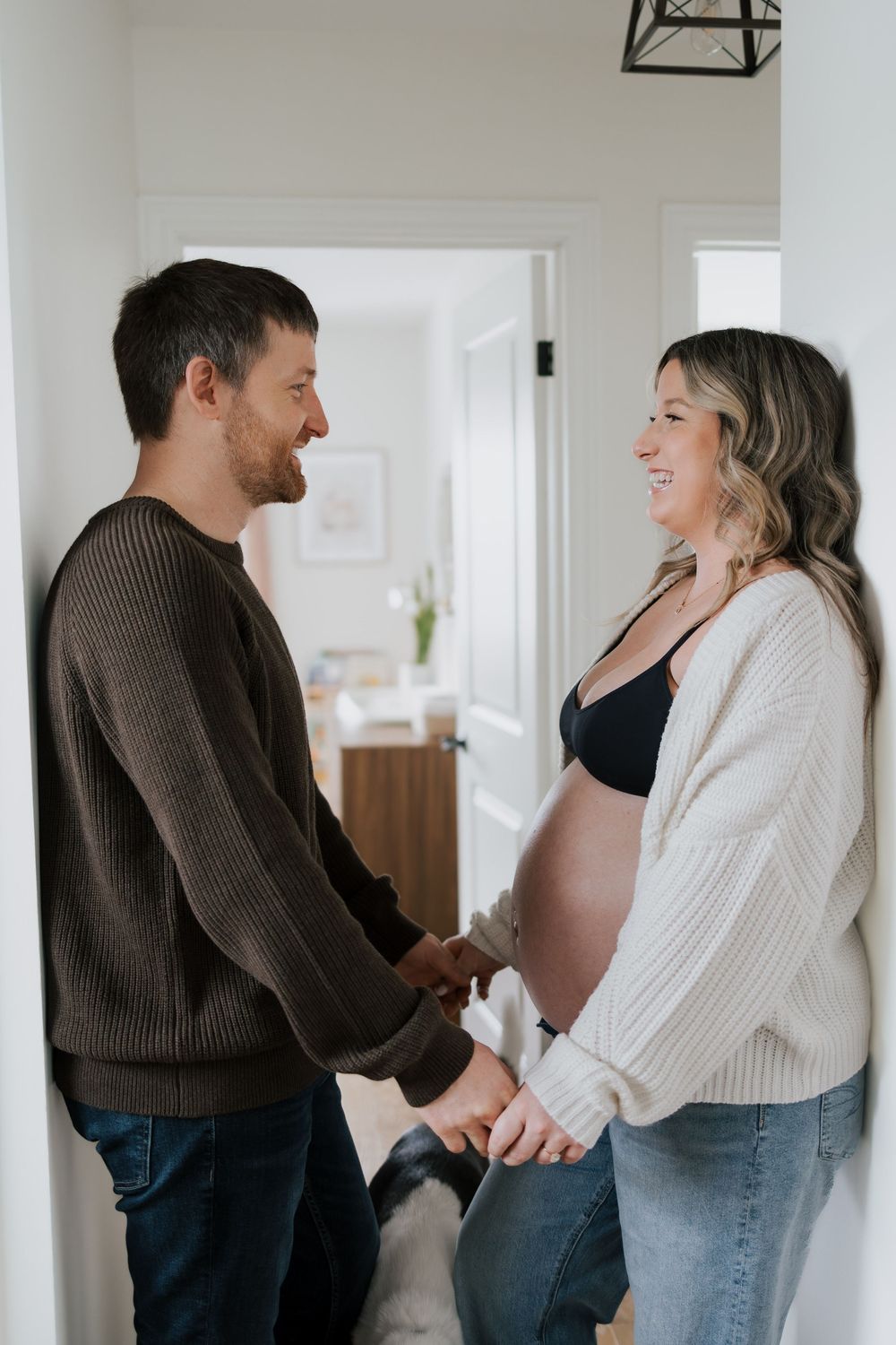 A happy couple holds hands in a hallway, the woman visibly pregnant, with their dog sitting nearby.