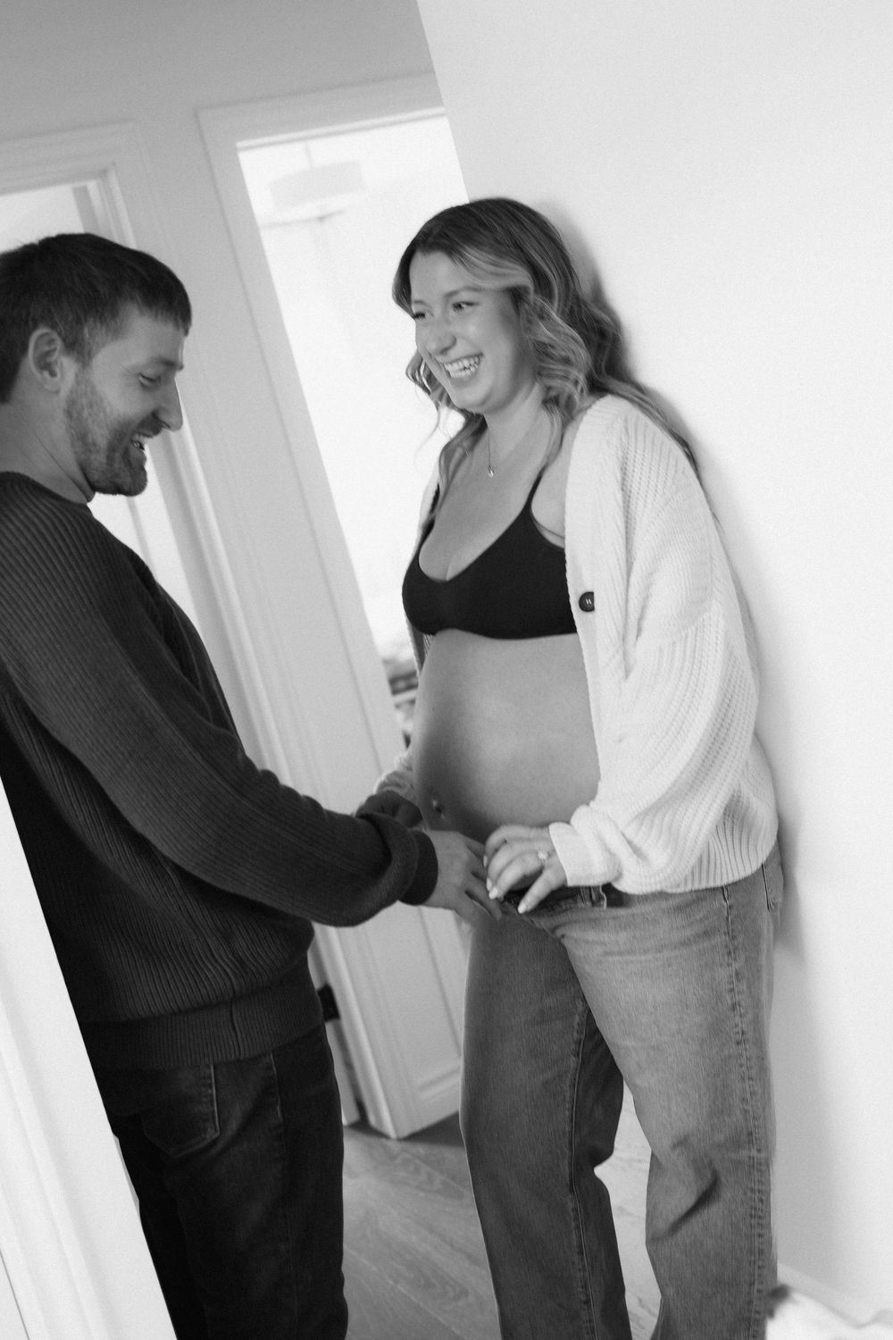 A black and white photo of a smiling pregnant woman and her partner holding hands in a bright hallway.