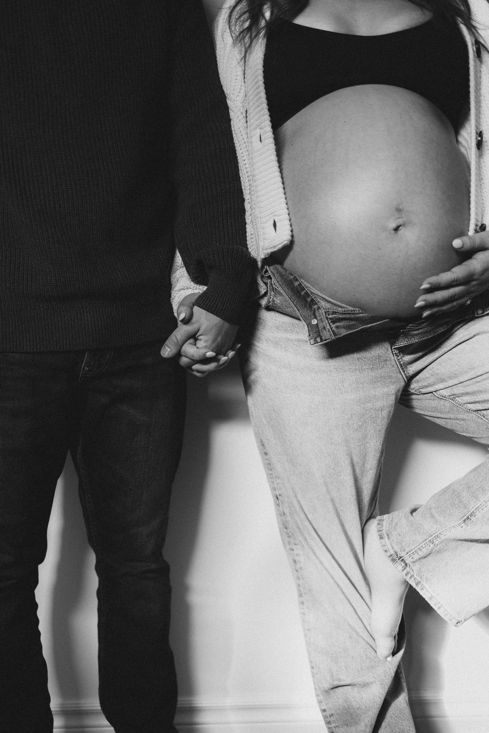 Black and white image of a pregnant woman and her partner holding hands against a white wall.