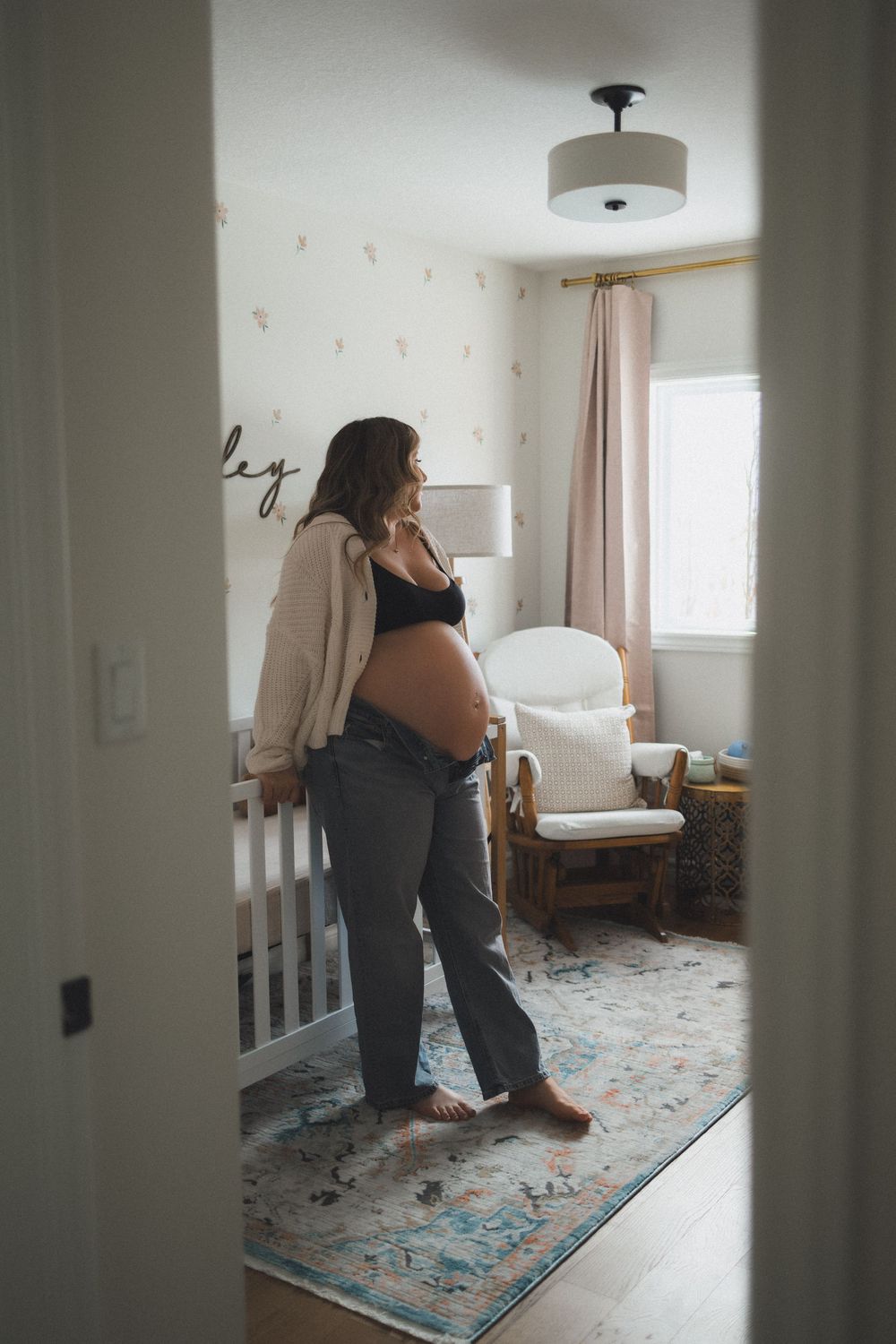 Pregnant woman in white cardigan and jeans standing in softly decorated baby nursery.
