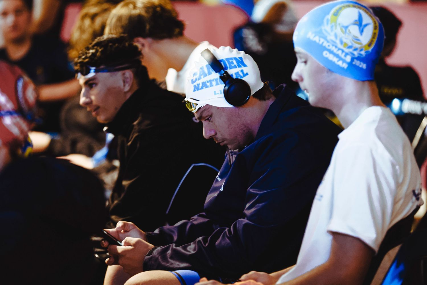 Female swimmers in caps and goggles stand at starting blocks during a competitive swim meet outdoors.