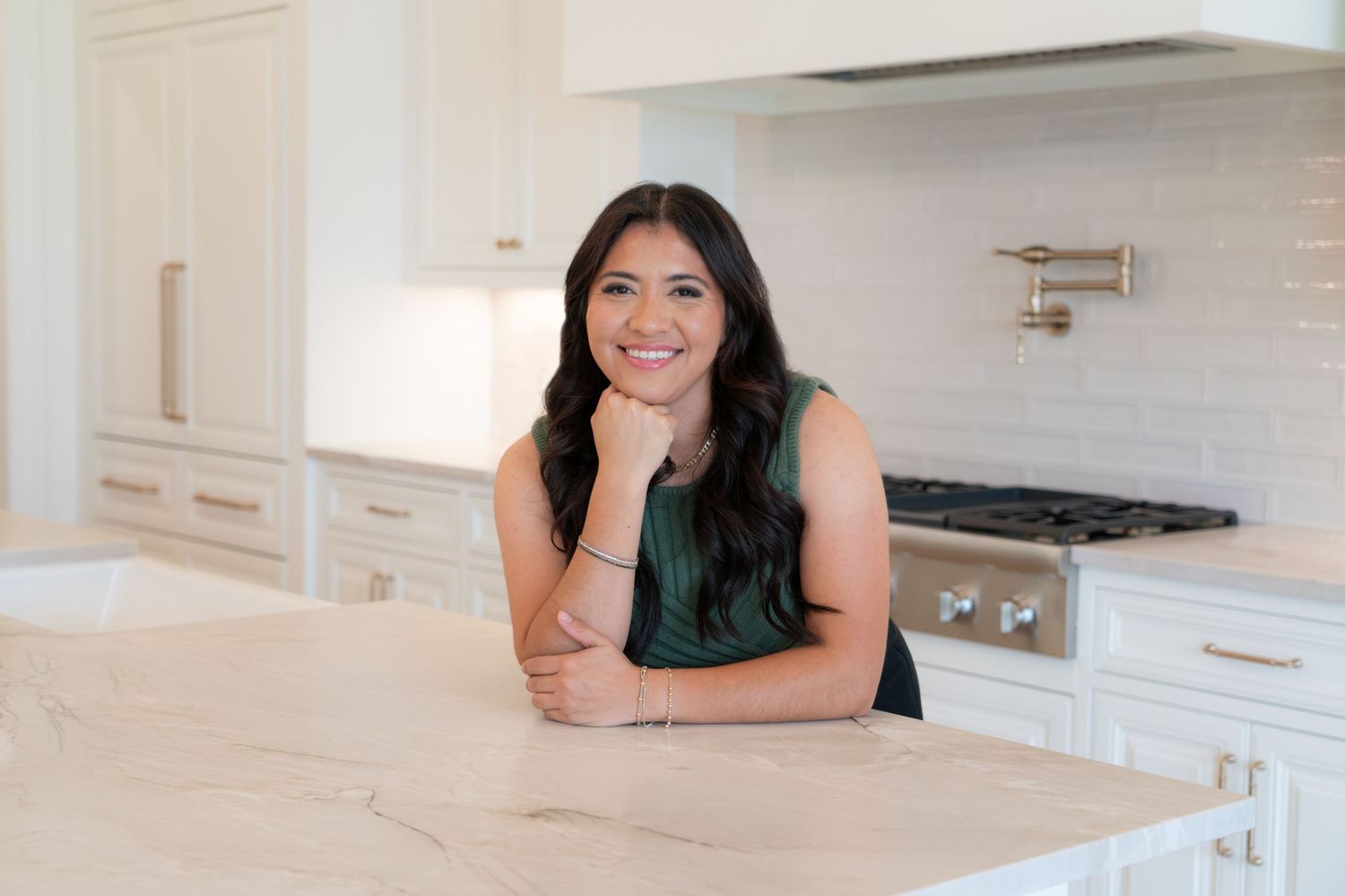 A smiling woman leans on a white marble kitchen island surrounded by elegant white cabinetry and a stainless steel stove.