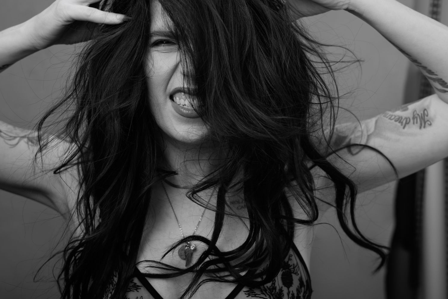 Black and white photo of a woman with long dark hair, tattoos on her arms, posing dramatically with hands in hair.