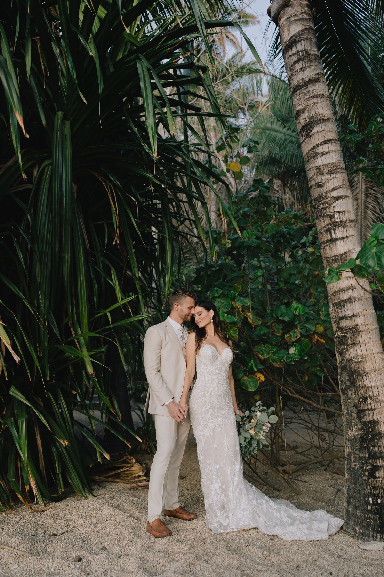 Bride and groom embrace among tropical palm trees in a lush garden, wearing a lace gown and tan suit.