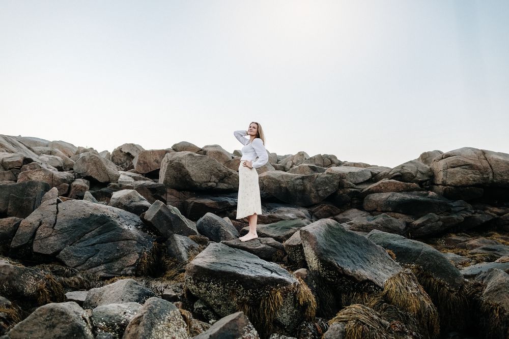 Blond girl wearing white blouse and yellow floral skirt stands posing on dark coastal rocks at Wingaersheek Beach in Gloucester, MA.